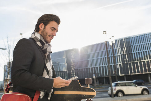 Deutschland, Frankfurt, Junger Geschäftsmann geht mit Skateboard durch die Stadt, benutzt Handy - UUF006335