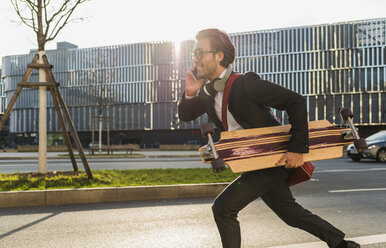 Germany, Frankfurt, Young businessman running with skateboard under his arm, using mobile phone - UUF006340