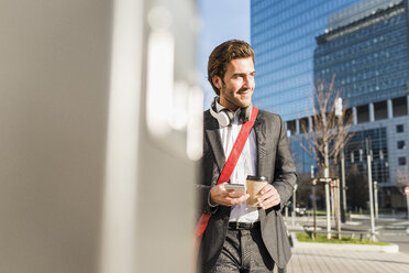 Deutschland, Frankfurt, Junger Geschäftsmann, der mit einer Tasse Kaffee durch die Stadt läuft und ein Mobiltelefon benutzt - UUF006344