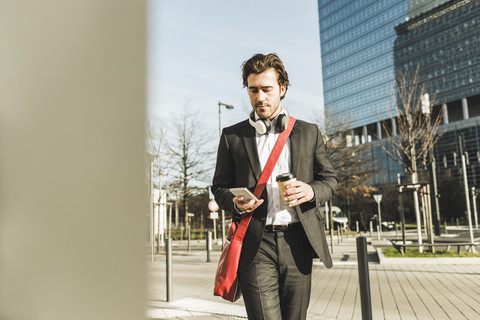 Deutschland, Frankfurt, Junger Geschäftsmann, der mit einer Tasse Kaffee durch die Stadt läuft und ein Mobiltelefon benutzt, lizenzfreies Stockfoto