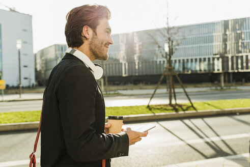 Germany, Frankfurt, Young businessman walking the city with cup of coffee, using mobile phone - UUF006347