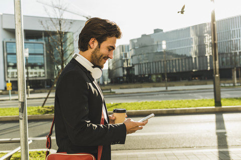 Deutschland, Frankfurt, Junger Geschäftsmann, der mit einer Tasse Kaffee durch die Stadt läuft und ein Mobiltelefon benutzt, lizenzfreies Stockfoto