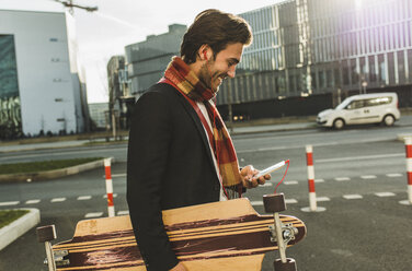 Germany, Frankfurt, Young businessman walking the city with skateboard, using mobile phone - UUF006350