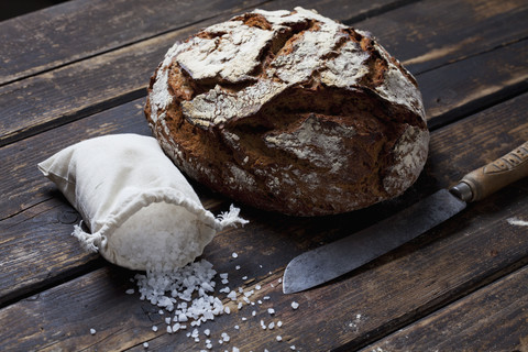 Knuspriges Brot, Brotmesser und Tütchen mit Salzkörnern auf dunklem Holz, lizenzfreies Stockfoto