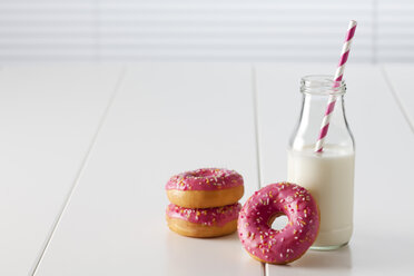 Glasflasche mit Milch und drei Krapfen mit rosa Zuckerguss auf weißem Grund - CSF027003