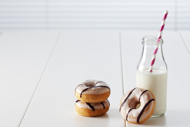 Glass bottle of milk and three doughnuts with icing on white ground - CSF027004