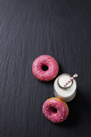 Glasflasche mit Milch und zwei Krapfen mit rosa Zuckerguss auf Schiefer, lizenzfreies Stockfoto