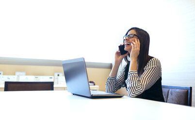 Laughing businesswoman sitting at desk telephoning with smartphone - MGOF001226