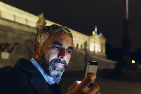Österreich, Wien, Porträt eines Mannes vor dem Parlamentsgebäude, der bei Nacht auf sein Smartphone schaut - AIF000206