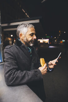 Austria, Vienna, man with Cheese Carniolan sausage and smartphone by night - AIF000210