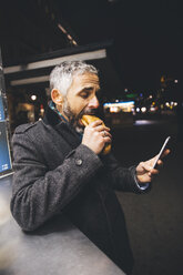 Austria, Vienna, man eating Cheese Carniolan sausage while looking at smartphone by night - AIF000211