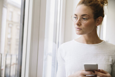 Junge Frau schaut von ihrem Handy auf, aus dem Fenster - MFF002587