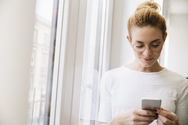 Young woman looking at her mobile phone, standing by window - MFF002588