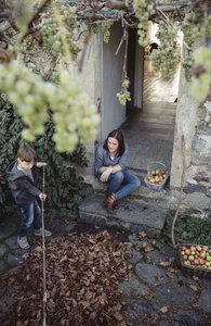 Spanien, Asturien, Frau sitzt am Eingang eines Landhauses und beobachtet ihren Sohn beim Pflücken von Herbstblättern - DAPF000002
