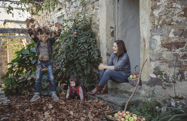 Spanien, Asturien, Frau sitzt am Eingang eines Landhauses und beobachtet ihre Kinder beim Spielen mit Herbstblättern - DAPF000003