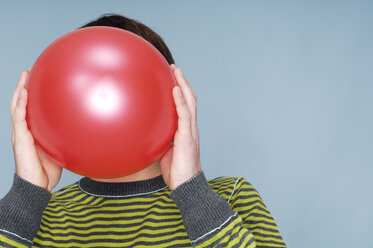 Teenage boy hiding face behind red balloon in front of blue background - GUFF000184