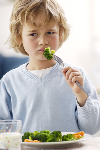 Portrait of unhappy blond boy eating broccoli stock photo