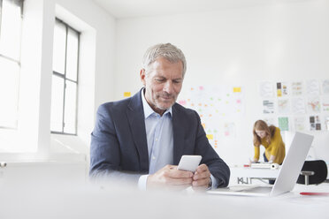 Geschäftsmann im Büro am Schreibtisch und schaut auf sein Handy - RBF004044