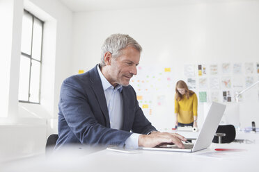 Smiling businessman in office at desk looking at laptop - RBF004045