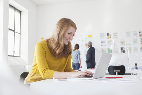 Lächelnde junge Frau im Büro am Schreibtisch mit Laptop, lizenzfreies Stockfoto