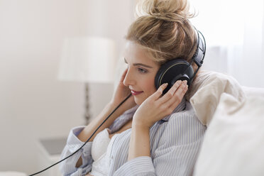 Portrait of smiling blond woman sitting on bed hearing music with headphones - SHKF000432