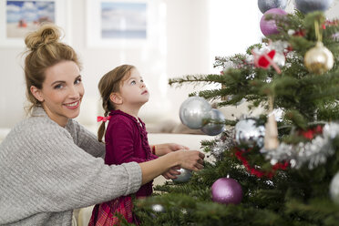Porträt einer lächelnden Frau beim Schmücken des Weihnachtsbaums mit ihrer kleinen Tochter im Wohnzimmer - SHKF000435