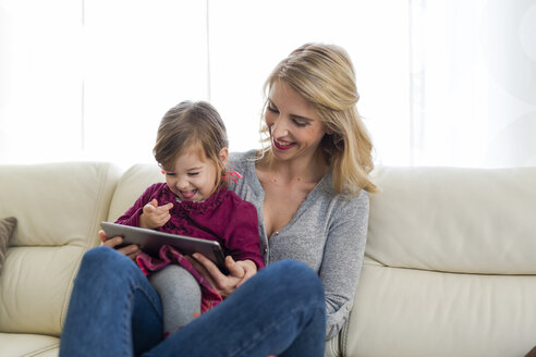 Mutter und ihre kleine Tochter sitzen auf der Couch im Wohnzimmer und schauen auf ein digitales Tablet - SHKF000438