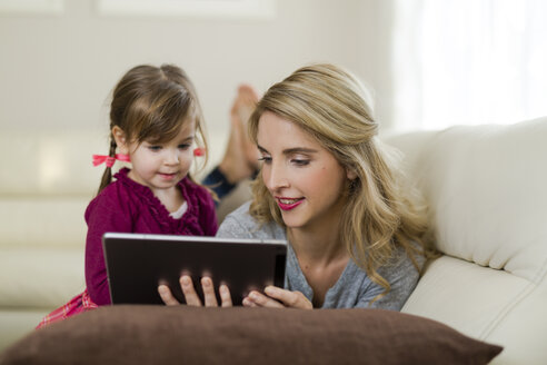 Mutter und ihre kleine Tochter sitzen zusammen auf der Couch im Wohnzimmer und schauen auf ein digitales Tablet - SHKF000441