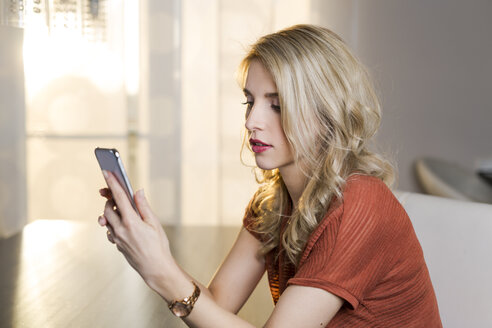 Blonde Frau sitzt auf der Couch im Wohnzimmer und schaut auf ihr Smartphone - SHKF000444