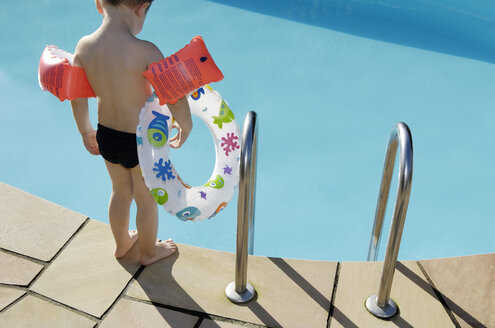 Back view of little boy with floating tire and water wings standing at pool edge - GUFF000229