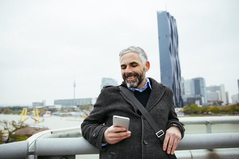 Austria, Vienna, smiling businessman standing on Reichsbruecke looking at his smartphone - AIF000218