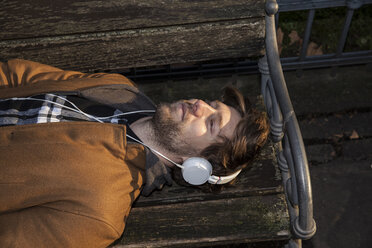 Porträt eines jungen Mannes, der auf einer Bank liegt und mit Kopfhörern Musik hört - FMKF002239