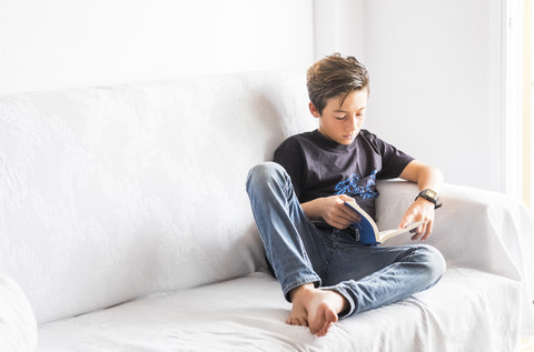 Junge sitzt auf einer weißen Couch und liest ein Buch, lizenzfreies Stockfoto