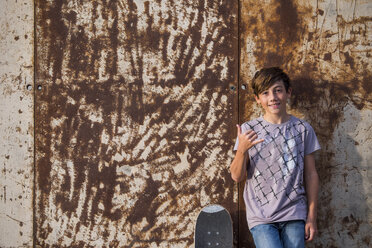 Portrait of boy leaning against rusty metal wall with his skateboard - SIPF000044