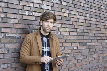 Porträt eines jungen Mannes mit digitalem Tablet vor einer Backsteinmauer - FMKF002245