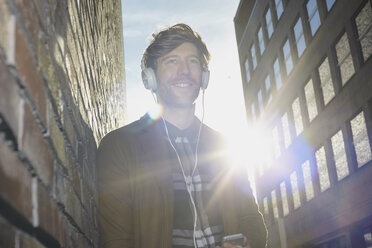 Smiling young man listening music with headphones at backlight - FMKF002248