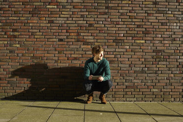 Junger Mann hockt mit digitalem Tablet vor einer Backsteinmauer - FMKF002254