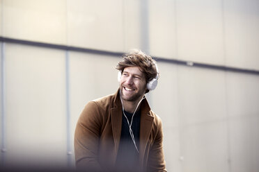 Smiling young man listening music with headphones at backlight - FMKF002259