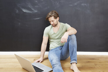 Young man sitting in front of chalkboard on the floor using laptop - FMKF002264