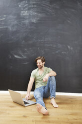 Porträt eines lächelnden jungen Mannes, der auf dem Boden vor einer Kreidetafel sitzt und einen Laptop benutzt - FMKF002265