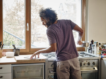 Young man standing at stove in kitchen checking digital tablet - RHF001248