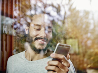Lächelnder junger Mann schaut auf ein Handy hinter einer Fensterscheibe - RHF001265