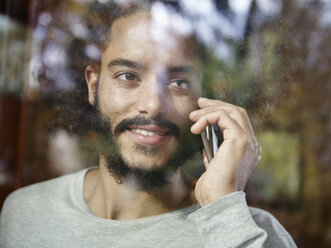 Smiling young man on cell phone behind windowpane - RHF001266