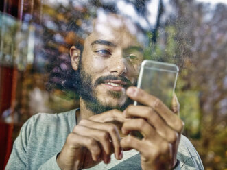 Junger Mann benutzt Mobiltelefon hinter einer Fensterscheibe - RHF001267