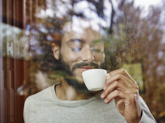 Smiling young man enjoying cup of coffee behind windowpane - RHF001268