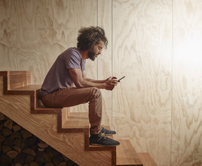 Junger Mann sitzt auf einer Holztreppe und schaut auf ein digitales Tablet - RHF001269