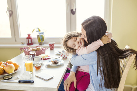 Porträt eines lächelnden kleinen Mädchens, das seine Mutter am Frühstückstisch umarmt, lizenzfreies Stockfoto