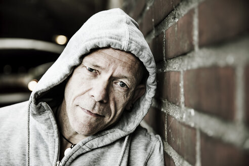 Portrait of man wearing grey hooded jacket leaning against brick wall - JATF000823