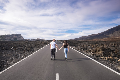 Spanien, Teneriffa, Rückenansicht eines Paares, das Hand in Hand auf einer leeren Straße im Teide-Nationalpark spazieren geht, lizenzfreies Stockfoto