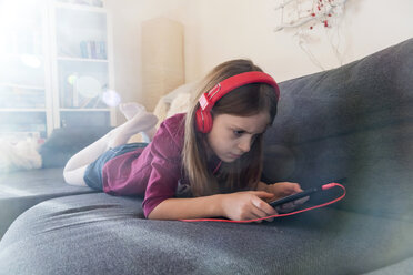Mädchen auf der Couch liegend und mit Kopfhörern und digitalem Tablet Musik hörend - SARF002460